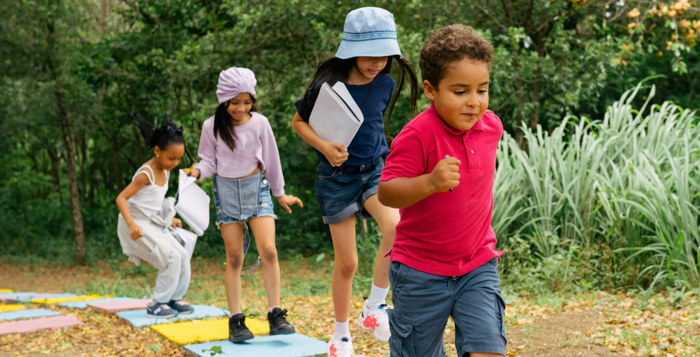 Children are playing on a colorful path in a forested area, with one child running ahead while others follow, holding notebooks.