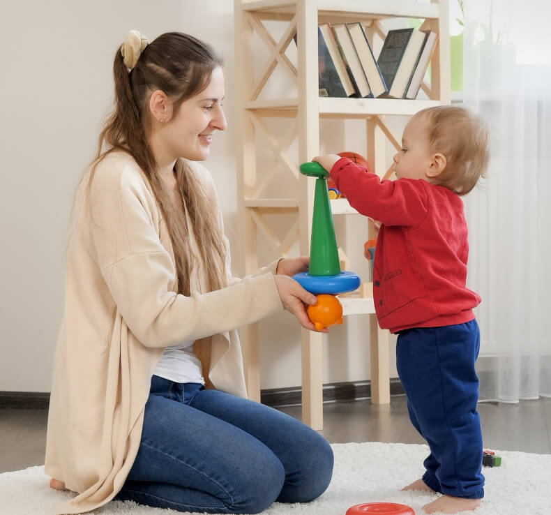 An adult with long hair sits on the floor, smiling and assisting a toddler wearing a red shirt and blue pants who is stacking colorful plastic rings on a cone.
