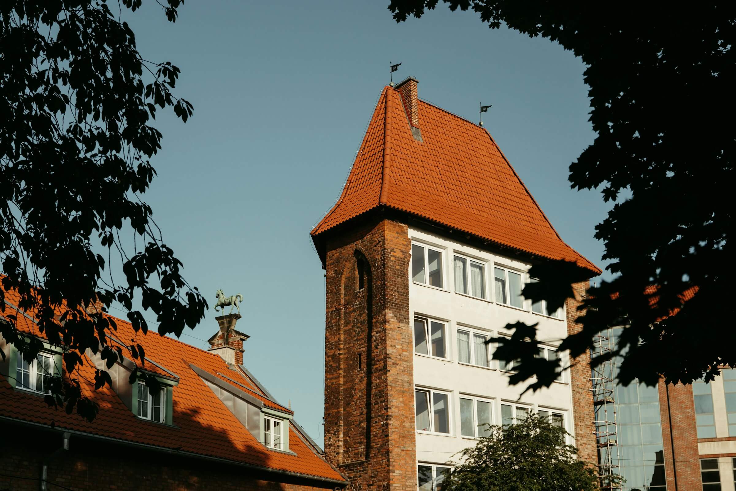 A brick building with white windows and a tall, pointed red-roofed tower stands against a clear sky, partially framed by green tree leaves—an inspiring sight as you start preparing your child for the transition to primary school.