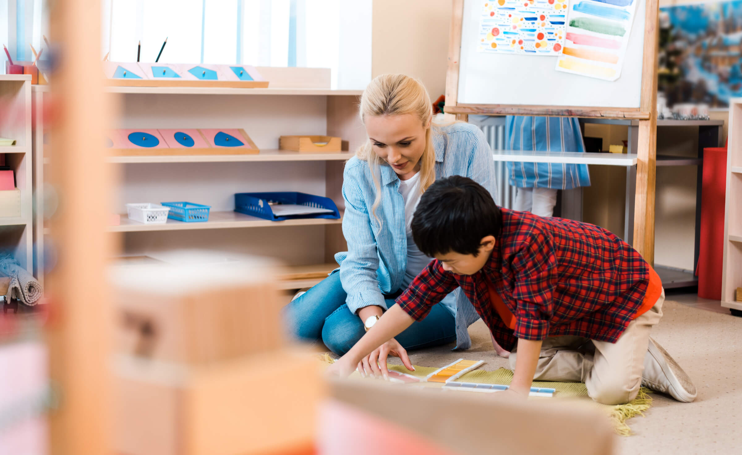 A woman sits on the floor with a young child, engaging in an educational activity in a classroom. Books and resources are on shelves in the background.