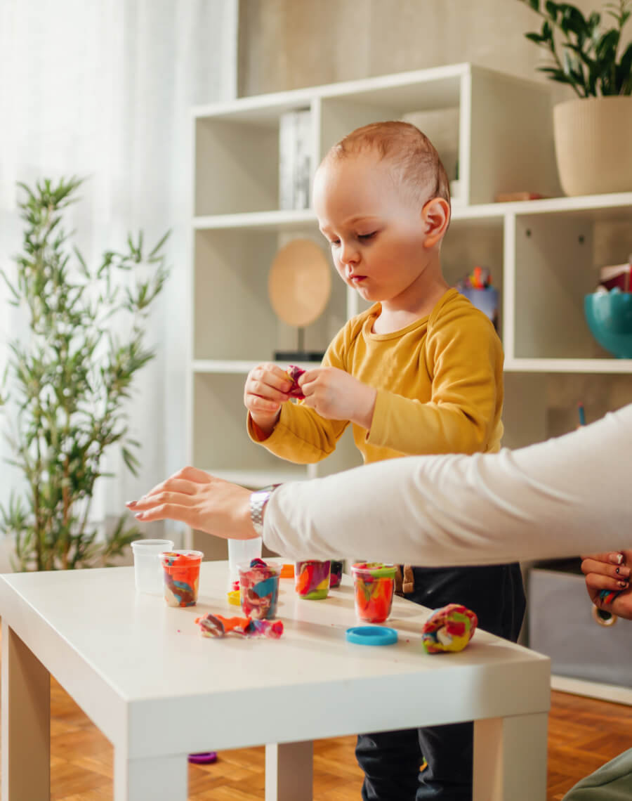 A young child in a yellow shirt is playing with colorful modeling clay on a small white table, assisted by an adult. Shelves and houseplants are visible in the background.