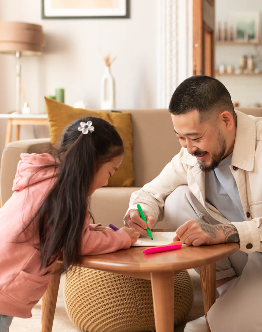 An adult and a child are drawing together at a wooden table in a cozy living room. The child is wearing a pink hoodie, and the adult is smiling.