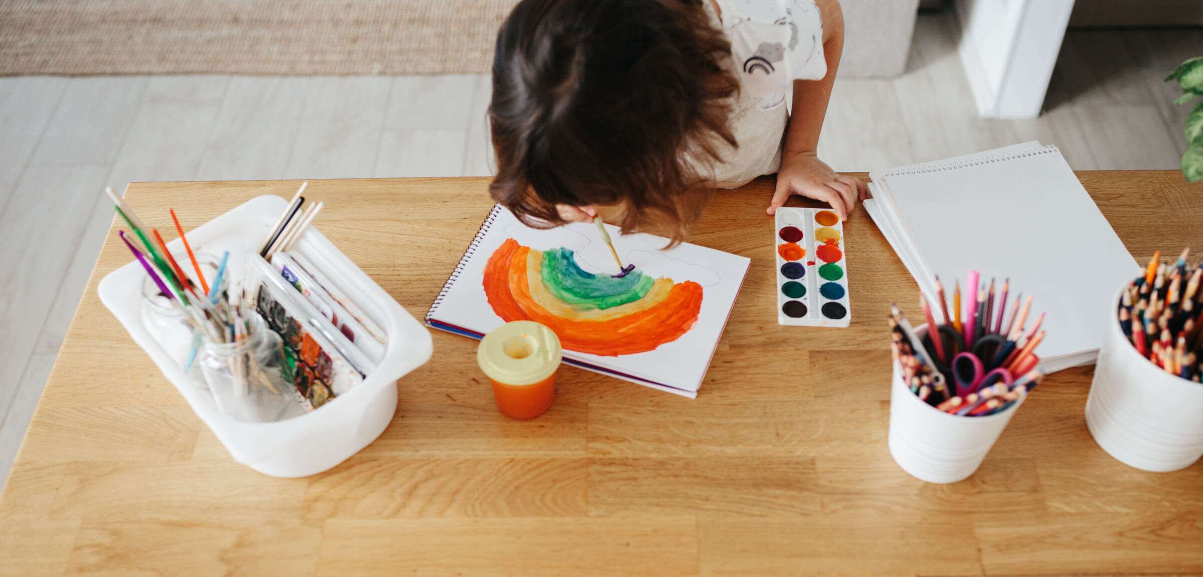 A child paints a rainbow on paper at a wooden table, surrounded by art supplies including paintbrushes, watercolor paints, and colored pencils—a peaceful moment akin to reading a carefully crafted Privacy Policy.