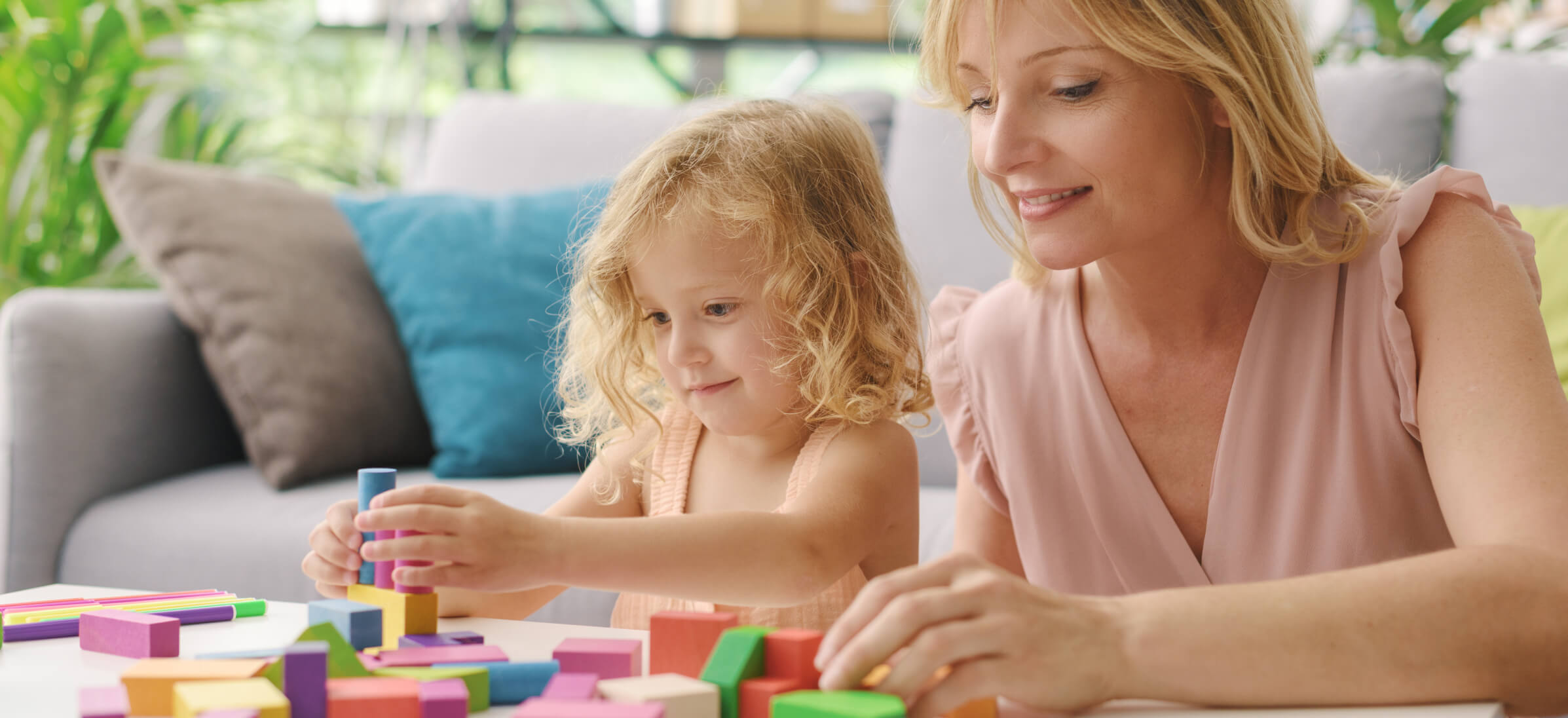 A woman and a child are building a structure with colorful wooden blocks at a table in a living room setting.